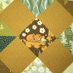 Op de afbeelding: Een patchworkquilt met een herhalend ruitpatroon. De quilt bevat verschillende stoffen met een natuurthema, waaronder een centrale ruit met een illustratie van een slapende vos. Andere ruiten tonen bloemen-, boom- en bladpatronen.