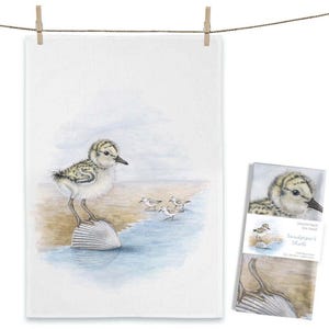 May include: A white tea towel featuring a watercolor illustration of a sandpiper chick standing on a seashell at the edge of the water. Three smaller sandpipers are in the background. The towel is hanging from a clothesline.