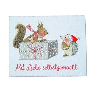 Könnte beinhalten: Ein rechteckiges Webetikett mit einer Stickerei eines braunen Eichhörnchens mit rot-weiß gestreiftem Schal auf einem grauen Geschenk mit weißen Punkten und roter Schleife, neben einem Igel mit roter Weihnachtsmütze und dem Schriftzug Mit Liebe selbstgemacht.