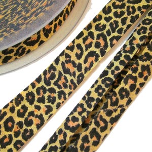 Könnte beinhalten: Eine Rolle Schrägband mit einem sich wiederholenden Leopardenmuster in Schwarz-, Braun- und Orangetönen. Das Stoffband ist an den Kanten gefaltet und zeigt ein durchgehendes Tiermuster über die gesamte Länge der Spule und des Bandes.