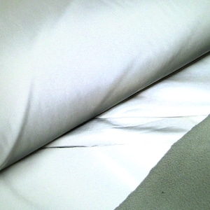 May include: Close-up of folded white fabric with a smooth texture. The fabric is layered, revealing a glimpse of a lighter, reflective material underneath. A section of textured, light green fabric is visible on the right side.