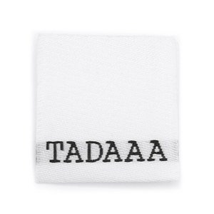 Könnte beinhalten: Ein weißes quadratisches Stoffetikett mit dem Wort "TADAAA" in Schwarz horizontal zentriert gedruckt. Das Etikett hat ein strukturiertes Aussehen und ist auf weißem Hintergrund isoliert.