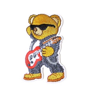 Könnte beinhalten: Ein gestickter Aufnäher, der einen goldbraunen Teddybären mit schwarzer Sonnenbrille und dunkelgrauer Jacke zeigt. Der Bär hält eine rot-weiße E-Gitarre und trägt hellbraune Stiefel, wobei die gesamte Figur von einem weißen Rand umgeben ist.