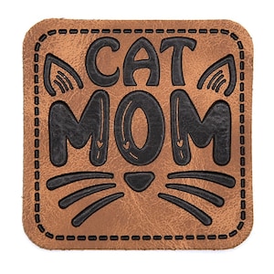 Könnte beinhalten: Ein quadratischer brauner Leder-Patch mit schwarzen Nähten und dem Text "CAT MOM" in einer stilisierten Schrift. Der Patch zeigt ein schwarzes Katzen Gesicht mit Schnurrhaaren und zwei Herzen über dem "O" in MOM.