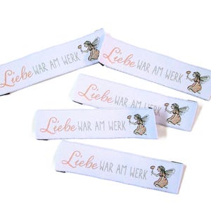 以下が含まれることがあります： ピンクとグレーの「Liebe WAR AM WERK」の文字が入った白い布製のラベル。各ラベルには、花を持っている妖精の小さなイラストが描かれています。ラベルは白い背景に配置されています。