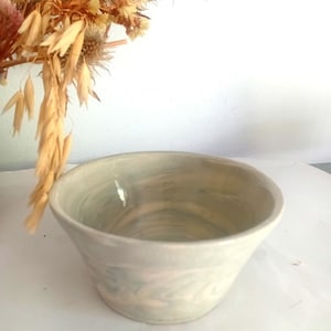May include: A ceramic bowl with a smooth, glossy finish featuring a marbled pattern of soft blue, white, and pale pink swirls. The bowl has a wide, open top that tapers down to a smaller, circular base, showcasing a rounded, flared rim.