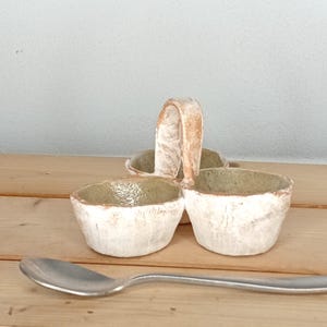 May include: A handmade ceramic condiment server with three small bowls and a handle. The bowls have a textured white exterior and a light green interior. A silver spoon rests in front of the server on a wooden surface.