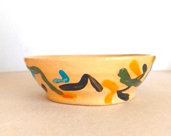 May include: A small, round, ceramic bowl with a light orange exterior. The bowl is decorated with colorful abstract shapes in blue, green, black, and orange. The bowl is likely handmade and suitable for serving small portions.