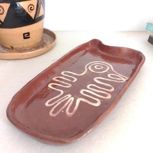 May include: A rectangular brown ceramic dish with a white stylized bird design. The dish is glazed and has a slightly raised rim.