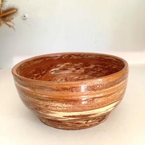 May include: A round ceramic bowl featuring a glossy finish with swirling patterns of terracotta, reddish-brown, and cream colors. The bowl has a smooth, curved interior and a slightly tapered base, showcasing a marbled effect across the exterior surface.