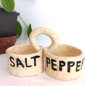 May include: Handmade ceramic salt and pepper holder with a handle. The beige holder has two compartments, each labeled in black with "SALT" and "PEPPER". The handle is a circular design. A plant is visible in the background.