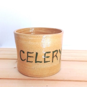 May include: A cylindrical, tan ceramic container with the word "CELERY" in dark green lettering. The container sits on a light-colored wooden surface, with a white background.
