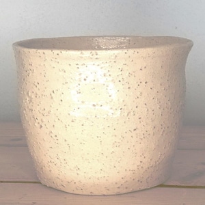 May include: A cylindrical ceramic planter with a wide, open top and a slightly flared rim. The exterior surface features a light cream-coloured glaze with a dense, speckled texture consisting of small, dark brown and grey flecks throughout.