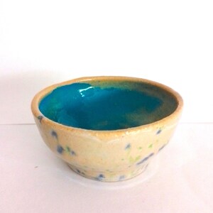 May include: A small, handmade ceramic bowl. The exterior is speckled beige, and the interior is a vibrant turquoise. The rim has a slightly uneven texture. The bowl is set against a white background.