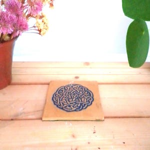 May include: A square, tan ceramic coaster with a blue Celtic knot design. The coaster is on a wooden surface, with a vase of pink flowers and a green leaf in the background.