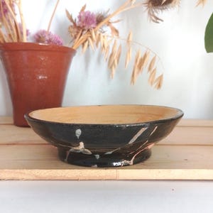 May include: A handmade ceramic bowl with a black exterior and a light tan interior. The bowl features white and pinkish-tan splatter designs. The bowl is on a wooden surface, with a terracotta vase and dried flowers in the background.