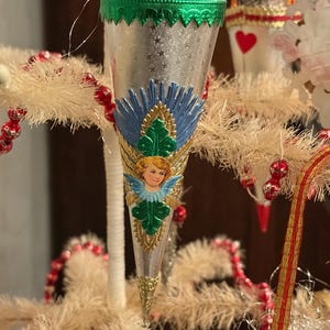 May include: A vintage Christmas ornament with a silver cone shape, adorned with a cherub, blue and gold accents, and a green scalloped trim. The ornament is part of a festive display with red beaded garland and white tinsel.