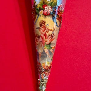 May include: A decorative cone-shaped ornament with a light blue background adorned with cherubic figures, flowers, and glitter. The ornament has a gold-colored tip and is likely used for decoration during the holidays.
