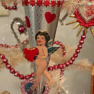 May include: A vintage-style Christmas tree ornament featuring a silver cone with red heart accents and a cherub holding a red heart. The ornament is decorated with red beaded garlands and star-shaped embellishments, evoking a romantic holiday theme.