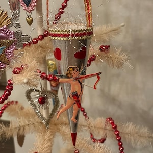 May include: A Valentine's Day-themed Christmas tree with red beaded garland and heart-shaped ornaments. A silver cone-shaped ornament with red hearts and a cupid figure is suspended from the tree. The tree is decorated with tinsel and other festive embellishments.