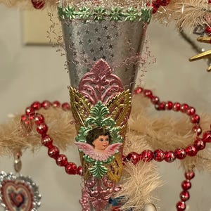 May include: A silver cone-shaped Christmas ornament with star patterns, green and pink accents, and a cherub image. The ornament is decorated with red beaded garland and other vintage ornaments.