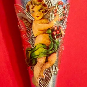 May include: Silver-toned decorative item featuring a cherub with wings, holding a wreath. The cherub is depicted in a vintage style with a green and yellow garment. The background is a vibrant red.