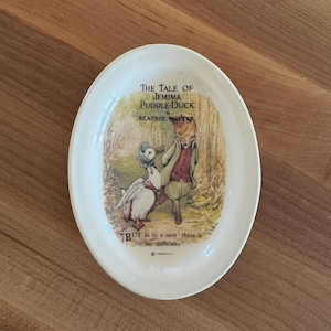 Puede incluir: Plato ovalado de cerámica crema con una ilustración de "The Tale of Jemima Puddle-Duck" de Beatrix Potter. La imagen muestra un pato y un zorro en un bosque. El plato incluye texto de la historia.
