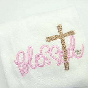 May include: White fabric with pink embroidery that says "blessed" and a brown cross above the word.
