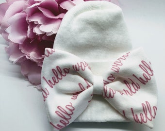 May include: White baby hat with a pink bow. The bow is made of fabric with the words "hello world" printed in pink.