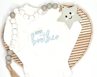 May include: White baby onesie with blue embroidery that reads "little brother". The onesie is on a wooden heart-shaped tray with a grey wooden star with a smiley face.