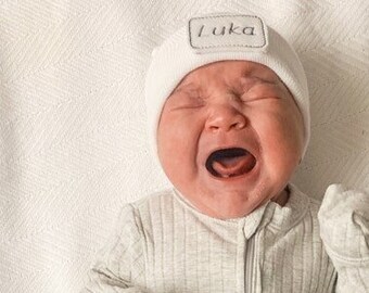 May include: A newborn baby wearing a white knit hat with the name "Luka" embroidered on it. The baby is wearing a gray ribbed onesie and is crying.