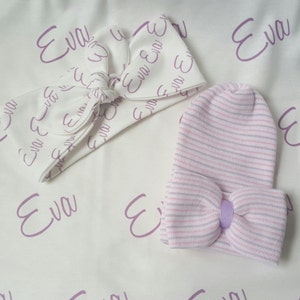 May include: A white headband with a bow and the name "Eva" printed in purple on it. A pink and white striped baby hat with a purple bow is also included.