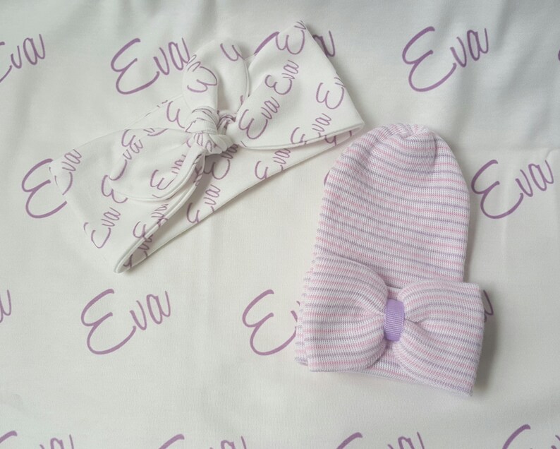 May include: A white headband with a bow and the name "Eva" printed in purple on it. A pink and white striped baby hat with a purple bow is also included.