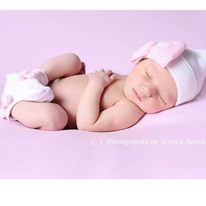 May include: A newborn baby girl sleeping on a pink background wearing a white and pink striped hat with a pink bow and white booties with pink accents.