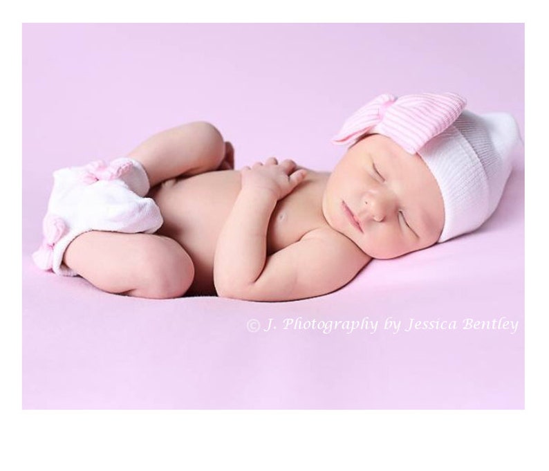 May include: A newborn baby girl sleeping on a pink background wearing a white and pink striped hat with a pink bow and white booties with pink accents.