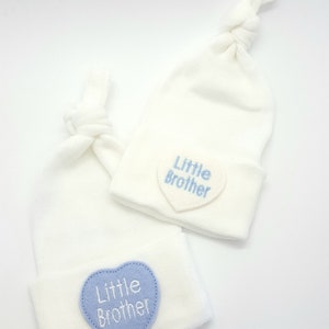 May include: Two white baby hats with blue heart-shaped patches that say "Little Brother" in white embroidery.