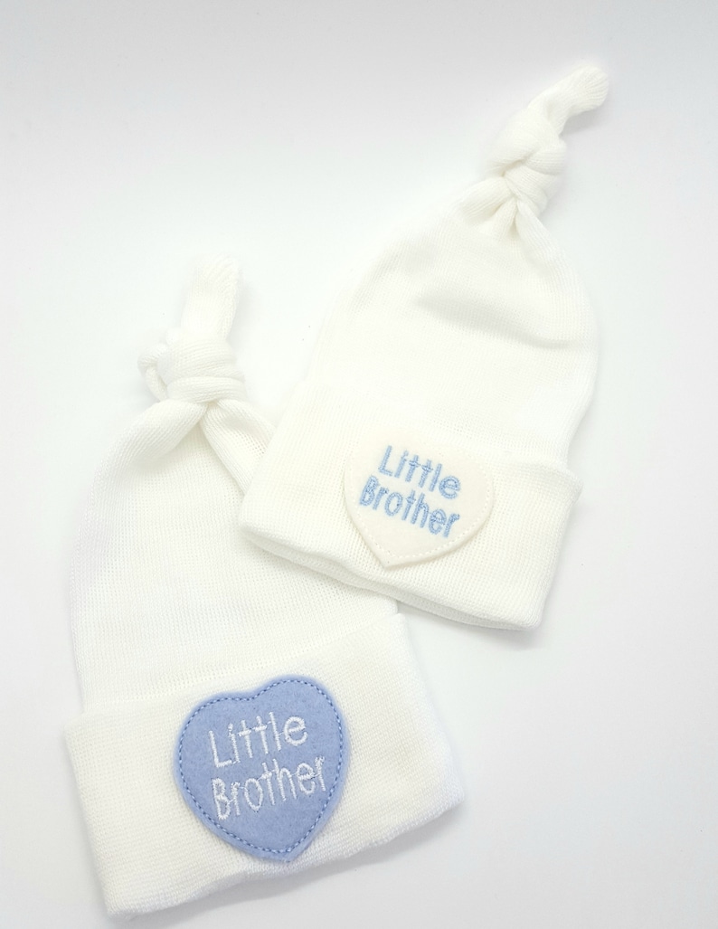 May include: Two white baby hats with blue heart-shaped patches that say "Little Brother" in white embroidery.