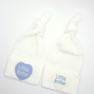 May include: Two white baby hats with blue and white heart-shaped patches that say "Little Brother".