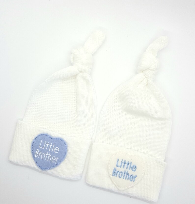 May include: Two white baby hats with blue and white heart-shaped patches that say "Little Brother".