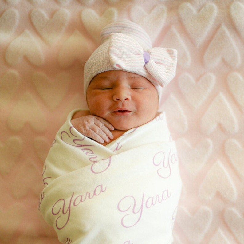 May include: A newborn baby girl is swaddled in a white blanket with the name "Yara" printed in purple. The baby is wearing a pink and white striped hat with a purple bow.