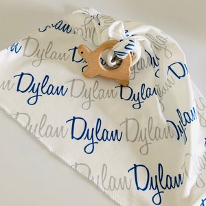 May include: A white baby blanket with the name "Dylan" printed in blue and gray script. A wooden turtle teether is on top of the blanket.