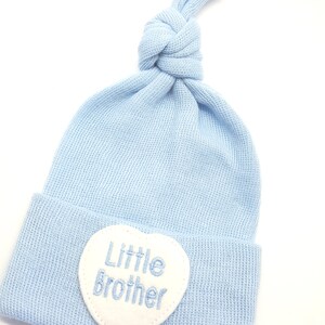May include: A light blue baby hat with a white heart-shaped patch that says "Little Brother" in blue embroidery.