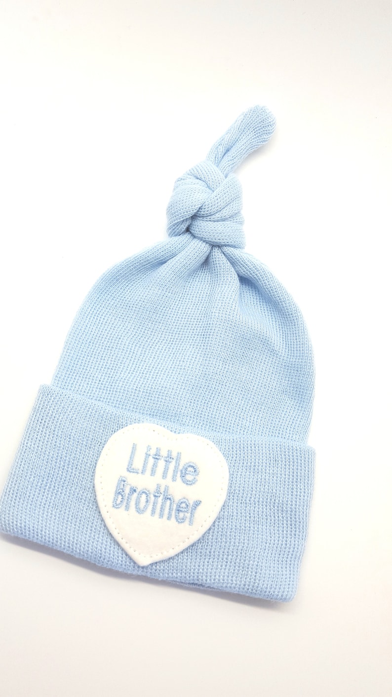 May include: A light blue baby hat with a white heart-shaped patch that says "Little Brother" in blue embroidery.