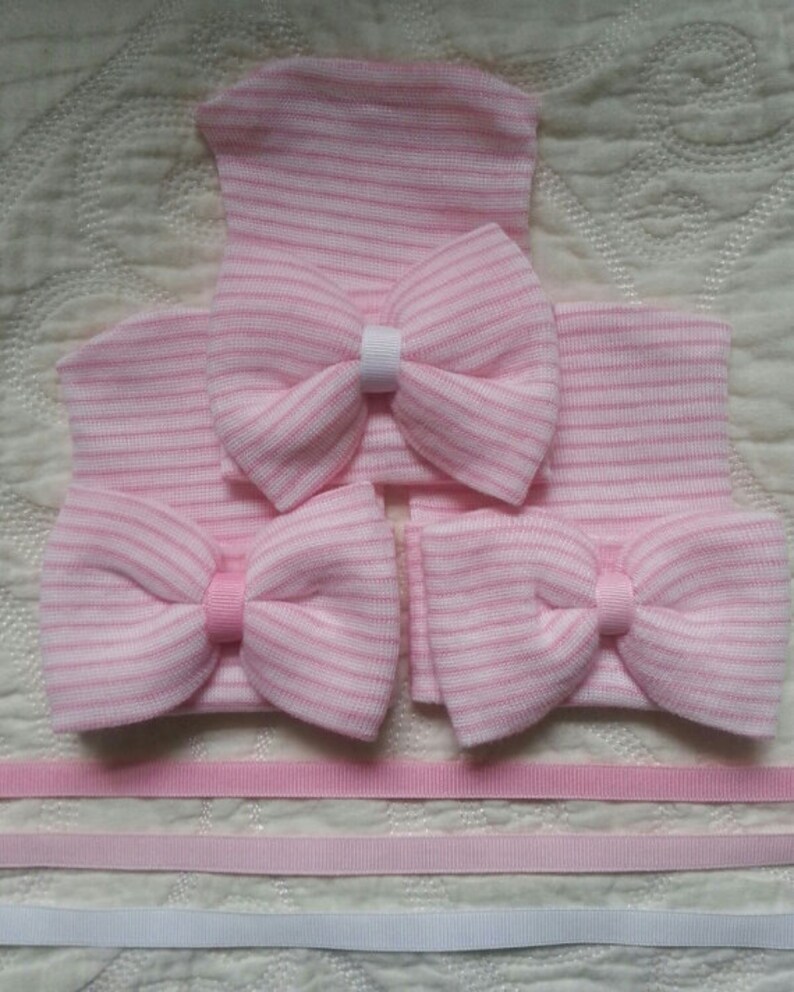 May include: Three pink and white striped baby hats with bows. The hats are made of soft knit fabric and have a simple design. The bows are made of the same fabric as the hats and are tied in a simple knot.