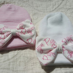 Op de afbeelding: Twee baby mutsen, een roze gestreept en een witte, elk versierd met een strik. De strikken zijn wit met de naam "Piper" in roze script. Deze newborn mutsen zijn ontworpen voor baby's.
