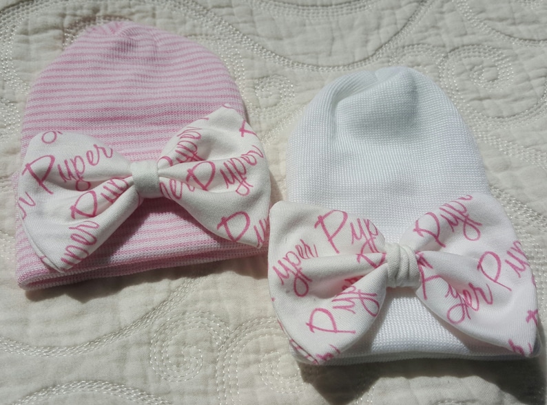 Op de afbeelding: Twee baby mutsen, een roze gestreept en een witte, elk versierd met een strik. De strikken zijn wit met de naam "Piper" in roze script. Deze newborn mutsen zijn ontworpen voor baby's.
