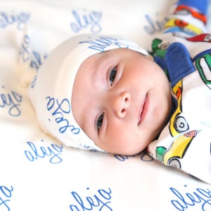 K&ouml;nnte beinhalten: Ein Baby tr&auml;gt eine wei&szlig;e M&uuml;tze mit blauer Aufschrift "Sofia" und einen wei&szlig;en und blauen Strampler mit einem bunten Auto-Druck. Das Baby liegt auf einer wei&szlig;en Decke mit blauer Aufschrift "aleijo", die sich wiederholt.