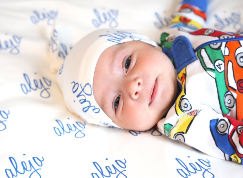 K&ouml;nnte beinhalten: Ein Baby tr&auml;gt eine wei&szlig;e M&uuml;tze mit blauer Aufschrift "Sofia" und einen wei&szlig;en und blauen Strampler mit einem bunten Auto-Druck. Das Baby liegt auf einer wei&szlig;en Decke mit blauer Aufschrift "aleijo", die sich wiederholt.