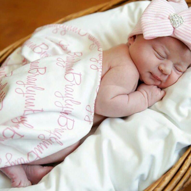 May include: A newborn baby girl sleeping in a white basket, wrapped in a white blanket with pink lettering that says "Brooklyn Baby". The baby is wearing a pink and white striped bow.