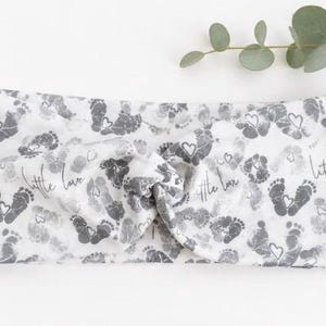 Pu&ograve; includere: Fascia per neonati bianca e grigia con un motivo ripetuto di impronte di piedi di bambino e cuori. Le parole "little love" sono stampate sulla fascia. Un rametto di foglie verdi &egrave; sullo sfondo.